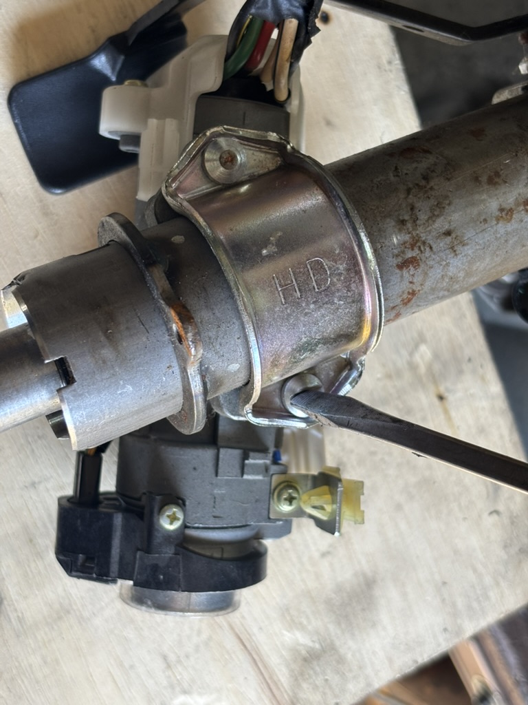 Flathead screwdriver removing a slotted steering column security bolt from ignition lock housing.